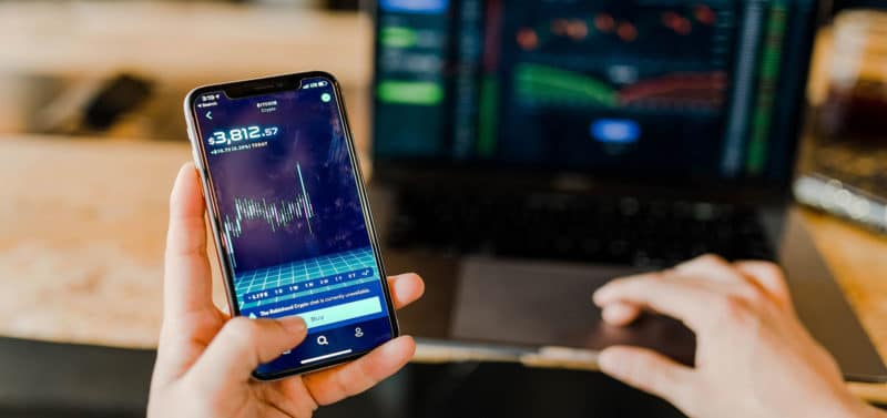 Stock image of person's hands, with one hand viewing the stock exchange on a smart phone and the other hand typing on a laptop computer.