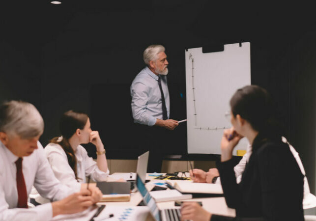 Group of coworkers in formal clothes gathering at table with papers and laptops while listening to male boss presenting business plan using flip chart in conference room